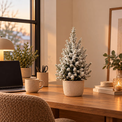 Kleine kerstboom met witte kunstsneeuw in een beige sierpot op een houten bureau naast een laptop en koffiemok.