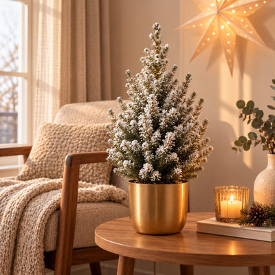 Kleine kerstboom met witte kunstsneeuw in een goudkleurige sierpot op een houten tafel naast een brandende kaars.